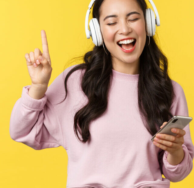 cropped-people-emotions-lifestyle-leisure-and-beauty-concept-carefree-happy-asian-woman-listening-music-in-wireless-headphones-holding-mobile-phone-singing-along-favorite-song-yellow-background.jpg
